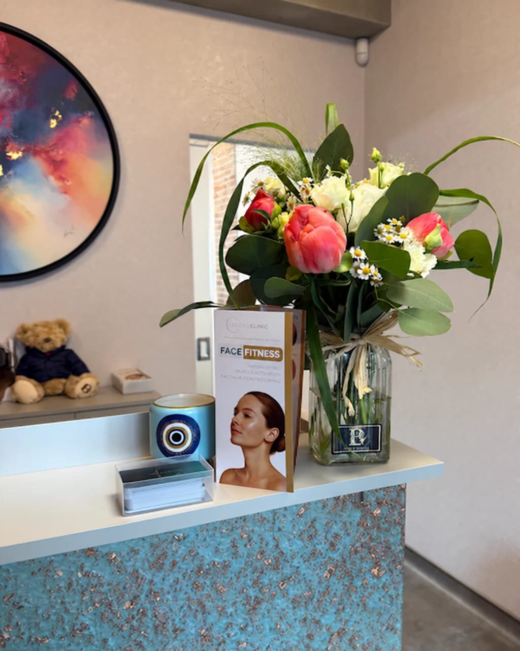 Reception desk at Levitas Clinic Esher with a vase of pink and white flowers, a Face Fitness leaflet and a round abstract painting on the wall