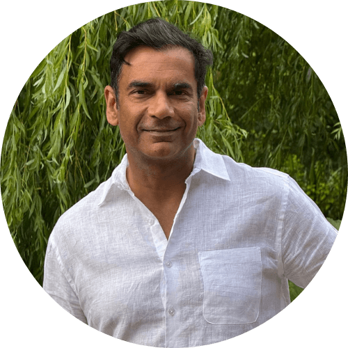 Dr Ash Kapoor smiling, wearing a white linen shirt, in front of a weeping willow