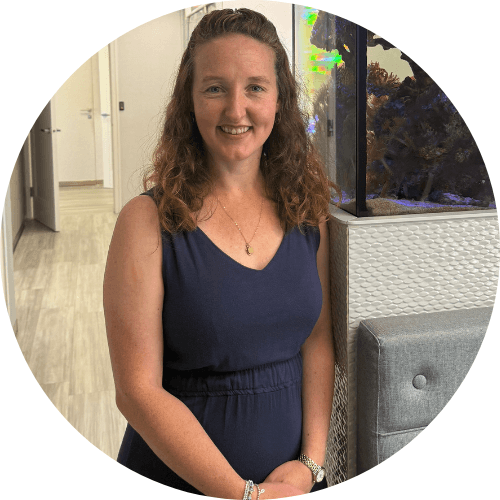 Dr Caitlin Scott smiling, with long curly hair, wearing a navy sleeveless dress in a clinic corridor