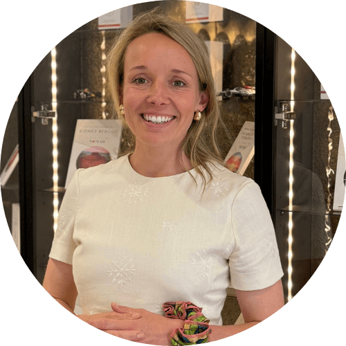 Dr Elizabeth Almas smiling, with fair hair, wearing a cream top in front of lit display shelves
