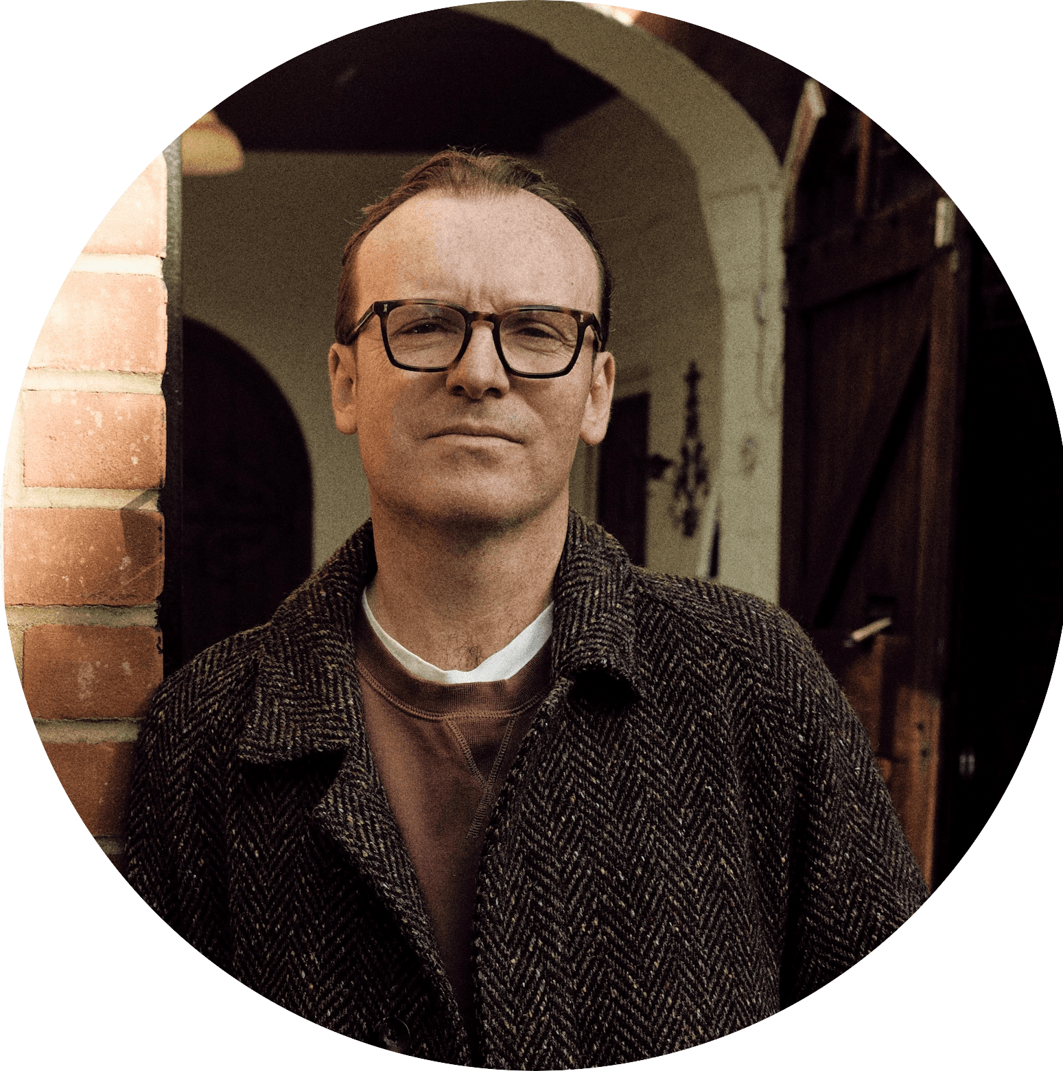 Tom Webb, wearing dark-rimmed glasses and a herringbone tweed coat, in front of a brick archway
